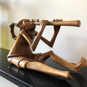 Może przedstawiać: Drewniana rzeźba przedstawiająca osobę grającą na flecie. Rzeźba jest rzeźbiona w stylizowanym, abstrakcyjnym stylu. Osoba siedzi ze skrzyżowanymi nogami i trzyma flet przy ustach. Rzeźba wykonana jest z jasnobrązowego drewna.