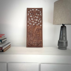 Peut inclure: Une sculpture murale en bois brun avec des sculptures complexes de deux personnages en vêtements traditionnels. Les personnages sont entourés de motifs floraux et d'un fond feuillu.