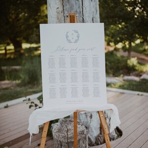 May include: A white wedding seating chart with a blue wreath and the initials "M & M" at the top. The chart lists guests' names by table number. The text "Please find your seat" is written in cursive at the top of the chart. The chart is displayed on a wooden easel.
