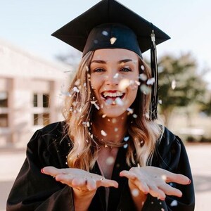 May include: A woman wearing a black graduation cap and gown smiles as she throws confetti into the air.