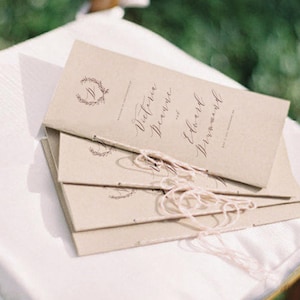 May include: Stack of brown paper wedding ceremony programmes with a white ribbon tied around them. The programmes have the names "Victoria & Edward" printed on the front.