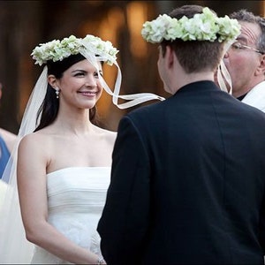 May include: A bride and groom exchange vows during an outdoor wedding ceremony. The bride is wearing a white strapless gown and a flower crown. The groom is wearing a black suit and a flower crown.