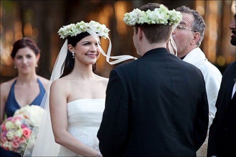 May include: A bride and groom exchange vows during an outdoor wedding ceremony. The bride is wearing a white strapless gown and a flower crown. The groom is wearing a black suit and a flower crown.