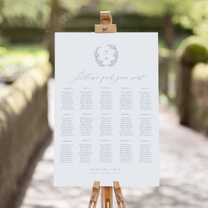 May include: A white wedding seating chart with a blue floral wreath and the initials "M & M" in the center. The chart lists guests by table number, with names printed in a light blue font. The text "Please find your seat" is written in cursive at the top of the chart.