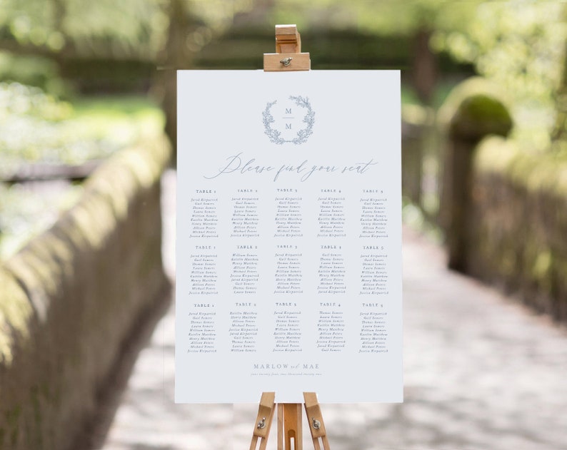 May include: A white wedding seating chart with a blue floral wreath and the initials "M & M" in the center. The chart lists guests by table number, with names printed in a light blue font. The text "Please find your seat" is written in cursive at the top of the chart.