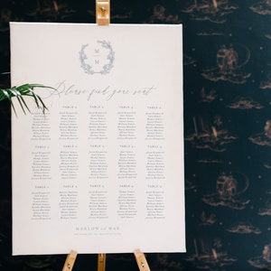 May include: A white seating chart with a blue and white floral wreath with the initials "M & M" at the top. The chart is titled "Please find your seat" and lists names under each table number.