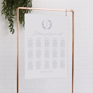 May include: A white wedding seating chart with a blue floral wreath and the initials "M & M" at the top. The chart lists guests' names by table number. The text "Please find your seat" is written in cursive. The chart is displayed on a copper stand.