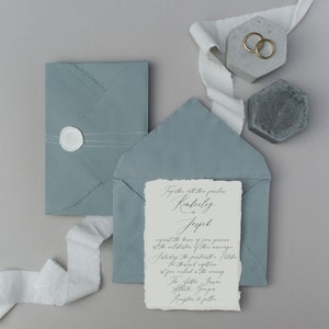 May include: A wedding invitation with a light blue envelope and a white card with calligraphy text. The text reads "Together with their families Kimberly + Jopel request the honor of your presence at the celebration of their marriage.  Saturday, the sixteenth of October.  The ceremony will take place at the venue.  The After, Lawn Reception to follow."