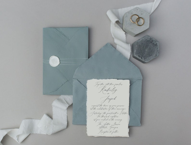 May include: A wedding invitation with a light blue envelope and a white card with calligraphy text. The text reads "Together with their families Kimberly + Jopel request the honor of your presence at the celebration of their marriage.  Saturday, the sixteenth of October.  The ceremony will take place at the venue.  The After, Lawn Reception to follow."