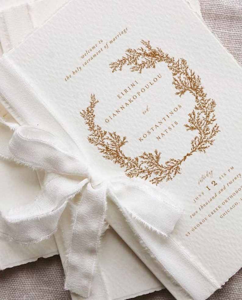 May include: A white wedding invitation with a gold floral wreath and the text "welcome to the holy sacrament of marriage" followed by the names "Eirini Giannakopoulou and Konstantinos Natsis" and the date "June 12 Six PM two thousand and twenty" and the location "St George Greek Orthodox Chicago".
