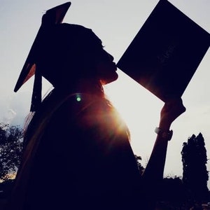 May include: Silhouette of a person wearing a graduation cap and gown, holding a diploma in front of a sunset sky.