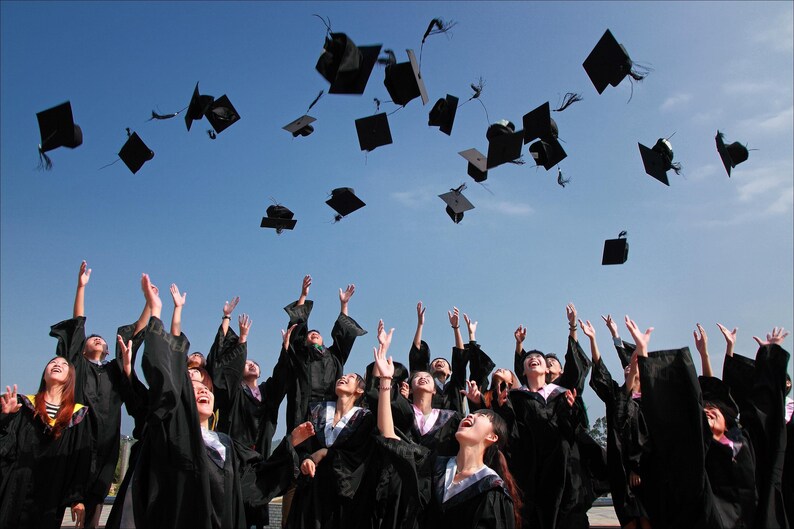 May include: A group of graduates in black caps and gowns throw their caps in the air during a graduation ceremony. The sky is blue and the graduates are smiling and excited.