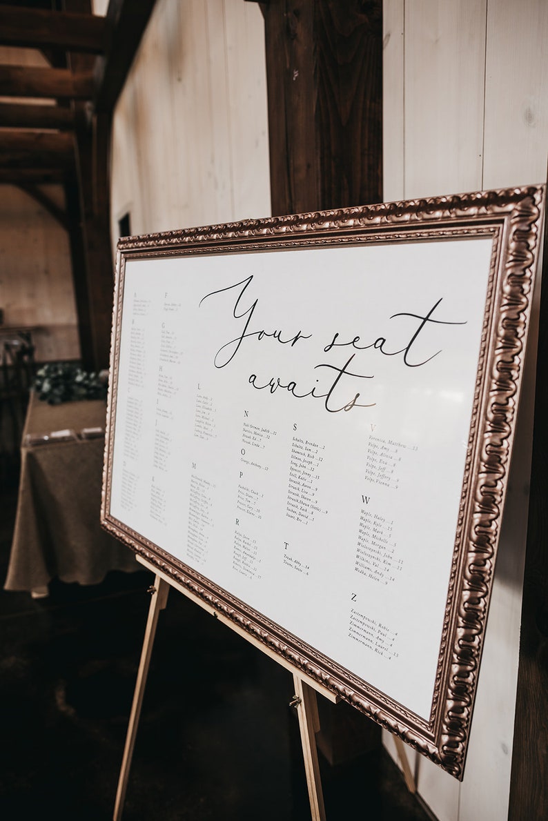 May include: A white framed seating chart with the text "Your seat awaits" at the top. The chart lists guest names alphabetically by last name with table numbers.