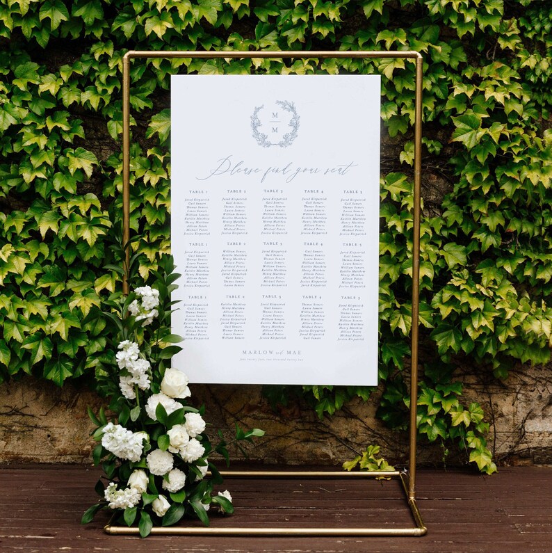 May include: A gold metal stand with a white poster displaying a wedding seating chart. The poster has a wreath with the letters "M & M" in the center and the text "Please find your seat". The poster lists the names of guests assigned to each table number.