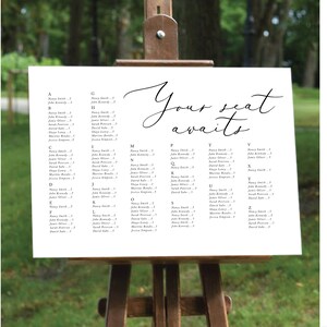 May include: A white seating chart with black text lists guest names and table numbers. The chart is on an easel and says "Your seat awaits".