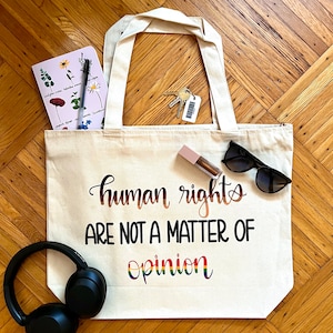 May include: A white canvas tote bag with the text "human rights are not a matter of opinion" in black lettering. The text "human rights" is in rainbow colors. The bag is on a wooden floor with other items, including a notebook, keys, sunglasses, and headphones.