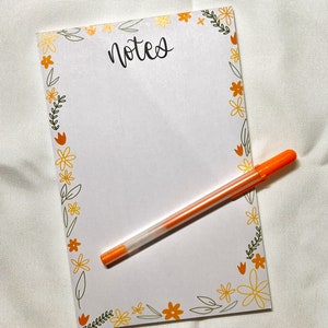Peut inclure: Un bloc-notes blanc avec le mot "notes" en écriture noire, entouré d'une bordure florale jaune, orange et verte. Un stylo orange est posé sur le bloc-notes. Le bloc-notes est conçu pour prendre des notes.