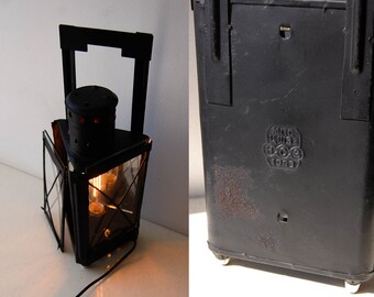 May include: A black metal lantern with a triangular frame, a top handle, and glass panels. The interior contains a glowing filament bulb. The exterior metal surface features a stamped emblem with the text "KPC" "1963" and shows signs of rust.