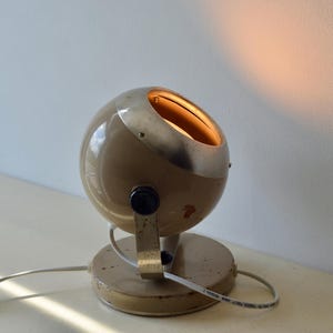 May include: A spherical table lamp with a beige metal casing and a silver-toned rim. The lamp features a circular base, a black adjustment knob on the side, and a white power cord. The interior of the sphere emits a warm, soft light.