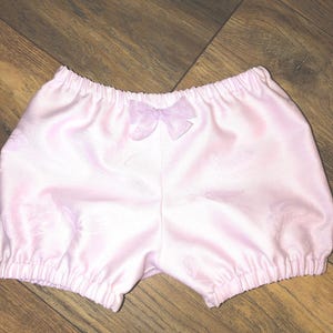 May include: Pink baby bloomers with a light purple bow. The bloomers have a gathered elastic waistband and a floral pattern.