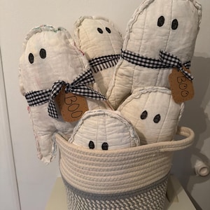 May include: A collection of five off-white fabric ghost plushies with black stitched eyes, each tied with a black and white gingham ribbon and a brown paper tag labeled BOO, all arranged inside a two-toned cream and gray woven rope basket.