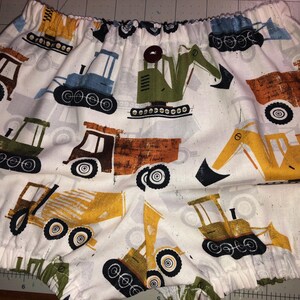 May include: White fabric with a repeating pattern of colorful construction vehicles, including excavators, dump trucks, and bulldozers. The vehicles are in shades of orange, green, blue, and yellow, with black wheels. The fabric is gathered at the top.