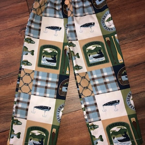 May include: Blue, green, and brown plaid pajama pants with a fishing theme. The pants have a variety of patches with images of fish, fishing lures, and fishing boats. The text "Fishing Northwest" is on two of the patches.