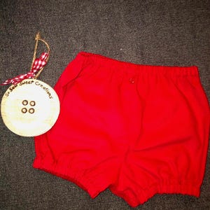 May include: Bright red baby bloomers with elastic waist and leg openings. A wooden ornament with the text "So Sew Sweet Creations" and a red and white checkered ribbon is next to the bloomers.