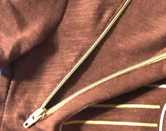 May include: A brown textured fabric garment featuring a diagonal gold-toned metal zipper closure. The material has a visible weave pattern, and a portion of a graphic design with horizontal white lines is visible at the bottom right corner.
