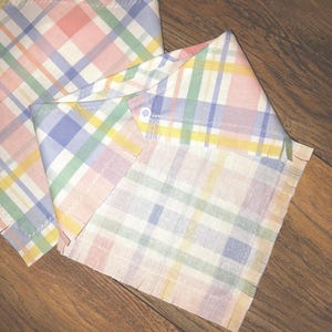 May include: A pink, blue, green, and yellow plaid bandana with frayed edges. The bandana is folded in half and has a button on the corner.
