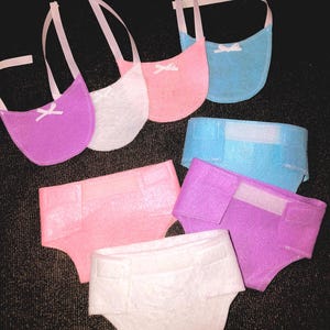 May include: Set of six felt doll accessories in pink, blue, purple, and white. The set includes three bibs with white ribbon ties and three diaper-style pants.