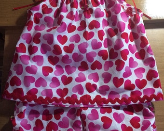 May include: A pink and white two-piece pajama set with a heart print. The top has thin red straps and a ruffled hem. The shorts have a scalloped hem.