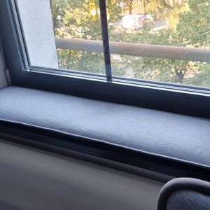 Puede incluir: Un coj&iacute;n gris para el asiento de la ventana est&aacute; colocado en el alf&eacute;izar de la ventana. El coj&iacute;n rectangular parece estar hecho de una tela suave. La ventana tiene un marco negro y da a &aacute;rboles y un edificio.