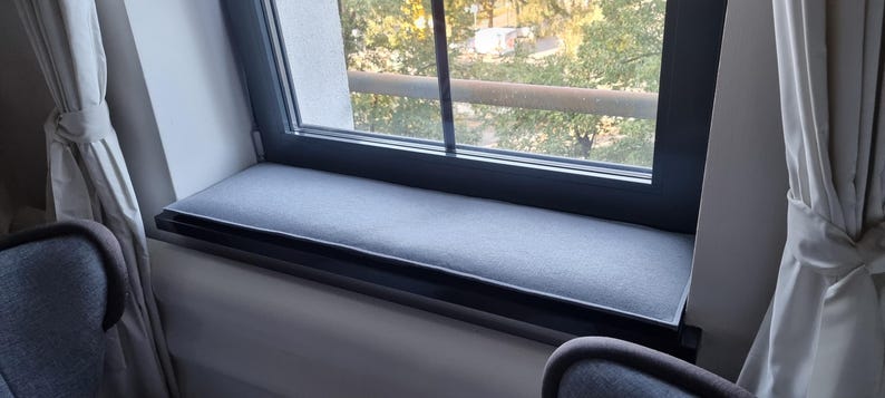 Puede incluir: Un coj&iacute;n gris para el asiento de la ventana est&aacute; colocado en el alf&eacute;izar de la ventana. El coj&iacute;n rectangular parece estar hecho de una tela suave. La ventana tiene un marco negro y da a &aacute;rboles y un edificio.
