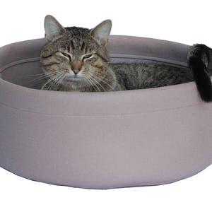 May include: A light pink cat bed with a gray tabby cat resting inside. The cat bed is made of soft, plush fabric and has a rounded shape.