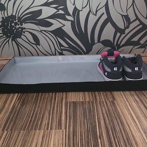Könnte beinhalten: Eine schwarze, rechteckige Schuhablage mit grauem Innenraum hält ein Paar rosa, schwarze und weiße High-Top-Sneaker. Die Sneaker haben ein Logo auf der Seite und stehen auf einem Holzboden.