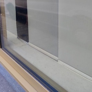 Puede incluir: Vista de cerca de una ventana con marco beige y cristal. La ventana est&aacute; parcialmente abierta, revelando una pared de color claro y un elemento vertical oscuro. El alf&eacute;izar de la ventana tambi&eacute;n es beige.