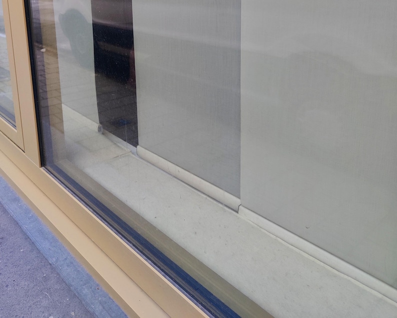 Puede incluir: Vista de cerca de una ventana con marco beige y cristal. La ventana est&aacute; parcialmente abierta, revelando una pared de color claro y un elemento vertical oscuro. El alf&eacute;izar de la ventana tambi&eacute;n es beige.