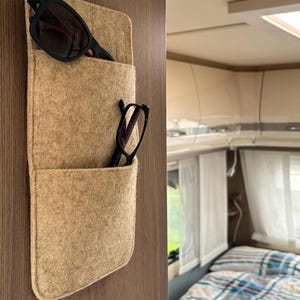 May include: A tan felt wall-mounted storage organizer with two stacked pockets attached to a wood-grain surface. The top pocket holds a pair of black sunglasses, and the bottom pocket holds a pair of black reading glasses with thin frames.