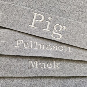 Puede incluir: Primer plano de tres tiras de fieltro gris con texto bordado en blanco. La tira superior dice "Pig", la tira del medio "Fellnasen" y la tira inferior "Muck". El fieltro tiene un aspecto texturizado.