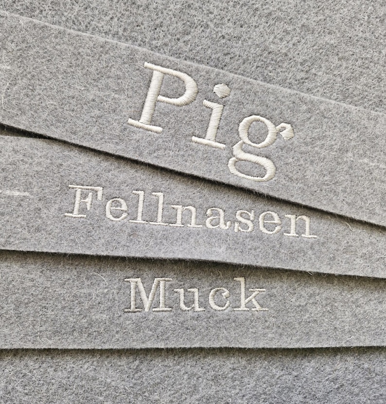 Puede incluir: Primer plano de tres tiras de fieltro gris con texto bordado en blanco. La tira superior dice "Pig", la tira del medio "Fellnasen" y la tira inferior "Muck". El fieltro tiene un aspecto texturizado.