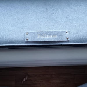 Puede incluir: Un coj&iacute;n tapizado azul claro con una etiqueta rectangular que dice "Bellmore". El coj&iacute;n forma parte de un mueble m&aacute;s grande, con un borde oscuro y una superficie de madera visible debajo.
