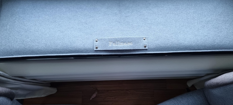 Puede incluir: Un coj&iacute;n tapizado azul claro con una etiqueta rectangular que dice "Bellmore". El coj&iacute;n forma parte de un mueble m&aacute;s grande, con un borde oscuro y una superficie de madera visible debajo.