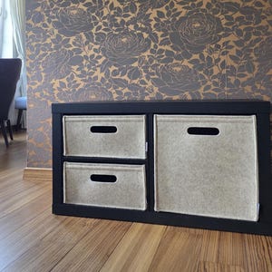 Op de afbeelding: Een zwarte opbergkast met drie beige stoffen bakken. De kast staat tegen een bloemenbehang in grijstinten en goud, en staat op een houten vloer.