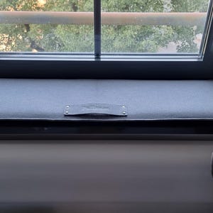 Puede incluir: Un asiento de ventana con un coj&iacute;n gris y una peque&ntilde;a etiqueta rectangular de cuero. El marco de la ventana es negro y da a una vista de &aacute;rboles. Dos sillones grises son visibles a ambos lados de la ventana.