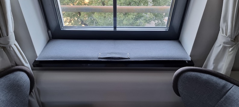 Puede incluir: Un asiento de ventana con un coj&iacute;n gris y una peque&ntilde;a etiqueta rectangular de cuero. El marco de la ventana es negro y da a una vista de &aacute;rboles. Dos sillones grises son visibles a ambos lados de la ventana.