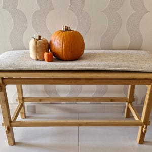 May include: A wooden bench with a light-colored upholstered seat. Three pumpkins of varying sizes and colors sit on the bench. The largest pumpkin is orange, the medium one is tan, and the smallest is orange.