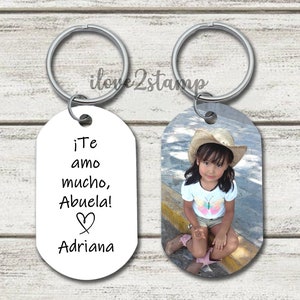 Peut inclure: Deux porte-cl&eacute;s argent&eacute;s avec une &eacute;tiquette ovale blanche. Une &eacute;tiquette a une photo d'une jeune fille portant un chapeau de paille et une chemise &agrave; imprim&eacute; papillon. L'autre &eacute;tiquette porte le texte "&iexcl;Te amo mucho, Abuela! Adriana" avec un symbole de c&oelig;ur.