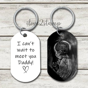Op de afbeelding: Twee zilveren sleutelhangers met een wit ovaal label waarop staat "Ik kan niet wachten om je te ontmoeten, papa!" en een zwart ovaal label met een zwart-witte echografie van een baby.