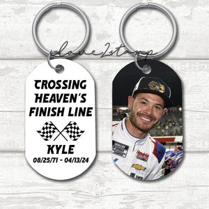 May include: Two oval-shaped keychains with a black and white checkered flag design. One keychain has a black background with a photo of a man wearing a racing uniform. The other keychain has a white background with the text "Crossing Heaven's Finish Line" and "Kyle 08/25/71 - 04/13/24".